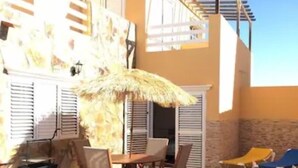 A sunny outdoor patio with a table and chairs, a thatched umbrella, and blue lounge chairs.
