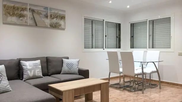 A modern living room with a grey sectional sofa, a wooden coffee table, and a glass-top dining table with four white chairs. There are two windows with shutters, and a large abstract painting hangs on the wall.