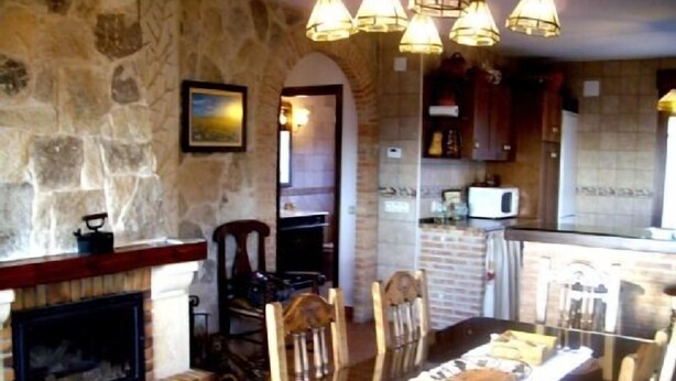 Un coin repas avec une cheminée, des chaises en bois, une table dressée avec de l’argenterie et des luminaires suspendus.
