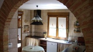 Une cuisine avec une arche en briques, une table ronde recouverte d’une nappe à motifs floraux, une lampe noire suspendue, une fenêtre avec des stores et un lave-vaisselle.