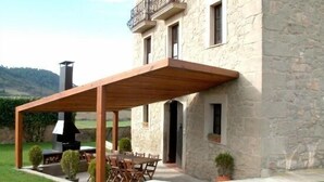 Ein Steinhaus mit einer hölzernen Pergola, Sitzgelegenheiten im Freien und einem Schornstein.
