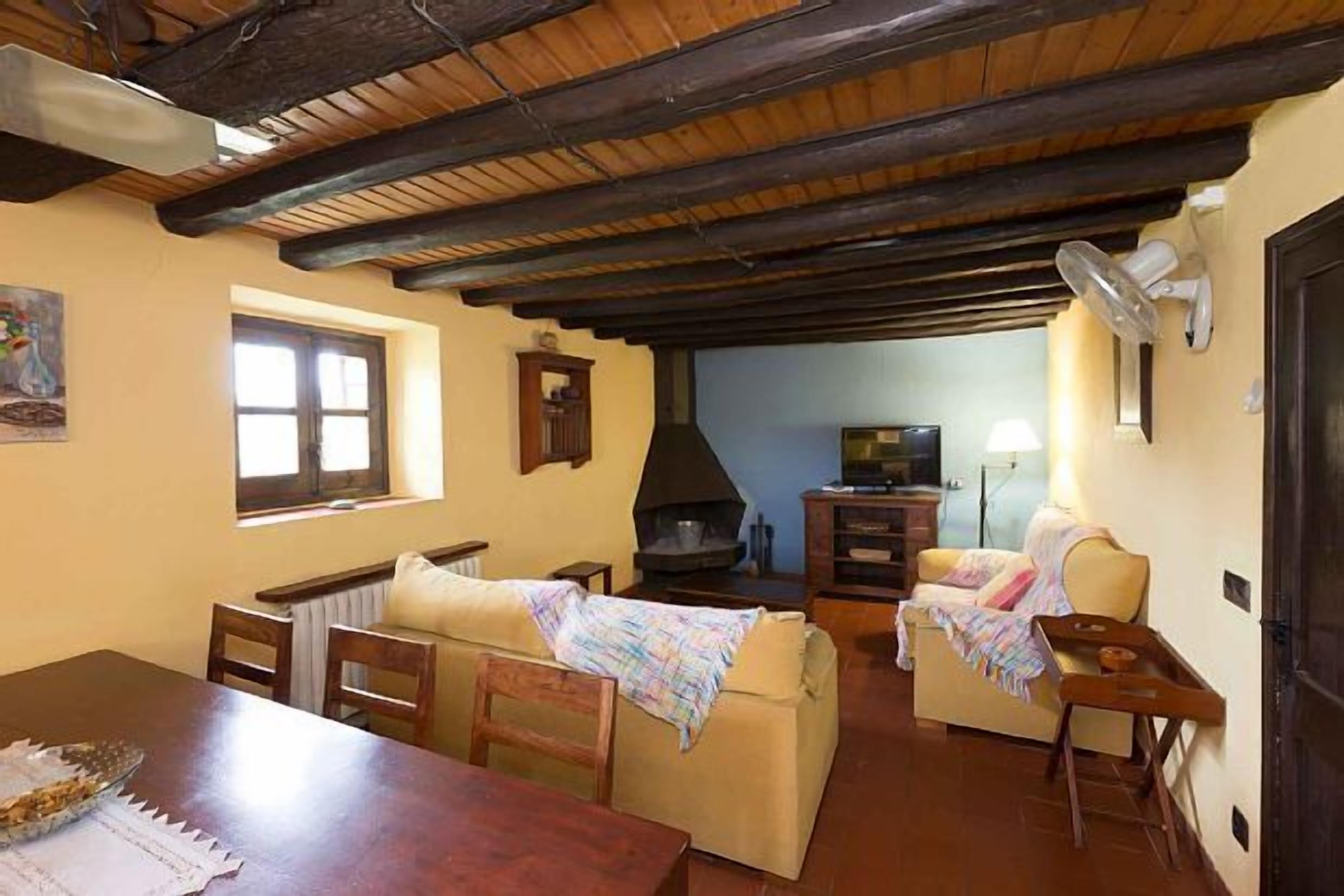 Ein gemütliches Wohnzimmer mit Holzbalkendecke, Kamin, Fernseher und Essbereich mit Tisch und Stühlen.