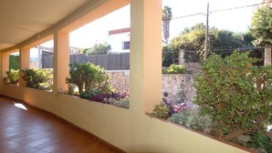 Un corredor cubierto con piso de baldosas, una serie de ventanas con plantas y una pared de piedra al fondo.