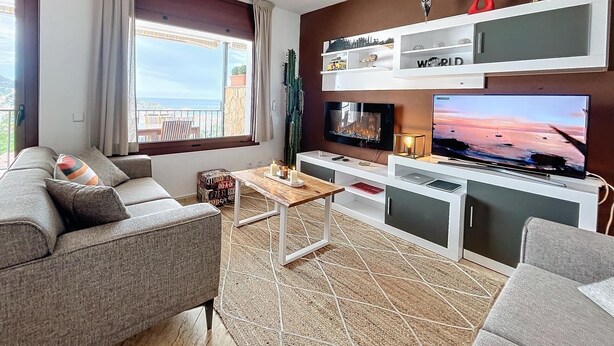 Una sala moderna con un sofá gris, una mesa de centro y un televisor montado en la pared. Se ve una ventana con vistas al mar y un balcón.