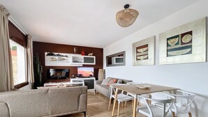 Una sala de estar moderna con un sofá gris, una mesa de comedor con sillas blancas, un televisor y una lámpara de techo.