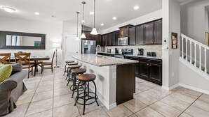 A modern kitchen with dark cabinets, a central island, bar stools, and a dining area with a table and chairs. - South Private Splash Pool • Lazy River • Resort Pool • 15 Minutes to Disney (Kissimmee)