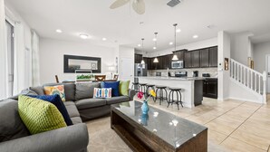 A modern living room with a gray sofa, a glass coffee table, and a kitchen area with dark cabinets and a white island. - South Private Splash Pool • Lazy River • Resort Pool • 15 Minutes to Disney (Kissimmee)