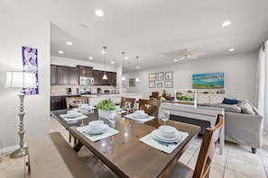 A modern kitchen with dark cabinets, a dining area with a long table set for four, and a ceiling fan.
