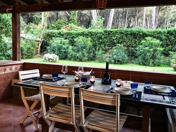 Un espace extérieur aménagé pour les repas, avec une table dressée pour quatre personnes, entourée de chaises, et une vue sur un jardin agrémenté d’une haie et d’arbres.