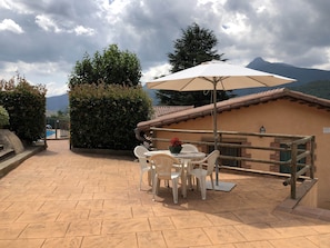 Un patio al aire libre con una mesa y sillas bajo un paraguas blanco, rodeado por una cerca de madera y con montañas al fondo.