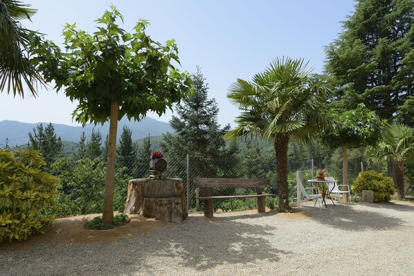 A serene outdoor seating area with a wooden bench and table, surrounded by lush greenery and a clear view of distant mountains.