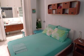 Een slaapkamer met een bed, een nachtkastje, een houten plank met boeken, een badkamer met een spiegel en een raam met gordijnen.