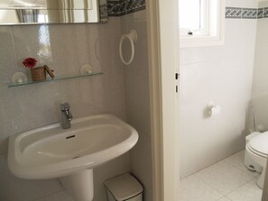 A bathroom with a white sink, a toilet, and a small trash bin. There is a glass shelf above the sink with a basket and a red flower.