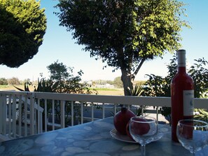 A table with a bottle of wine, two glasses, and a pomegranate on a terrace with a view of trees and a clear sky.