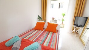 Una cama con ropa de cama a rayas y almohadas naranjas, un televisor sobre un mueble bajo y una ventana con cortinas.