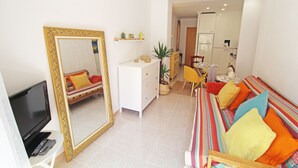 Una sala de estar con una televisión, un espejo grande, un sofá con cojines de colores, un aparador blanco y una planta en maceta.