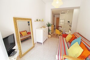 Un salon comprenant une télévision, un grand miroir, un canapé agrémenté de coussins colorés, une commode blanche et une plante en pot.