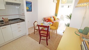Una cocina compacta con armarios blancos, una zona de comedor con una mesa y sillas amarillas, y una pequeña mesa con platos y un cactus.