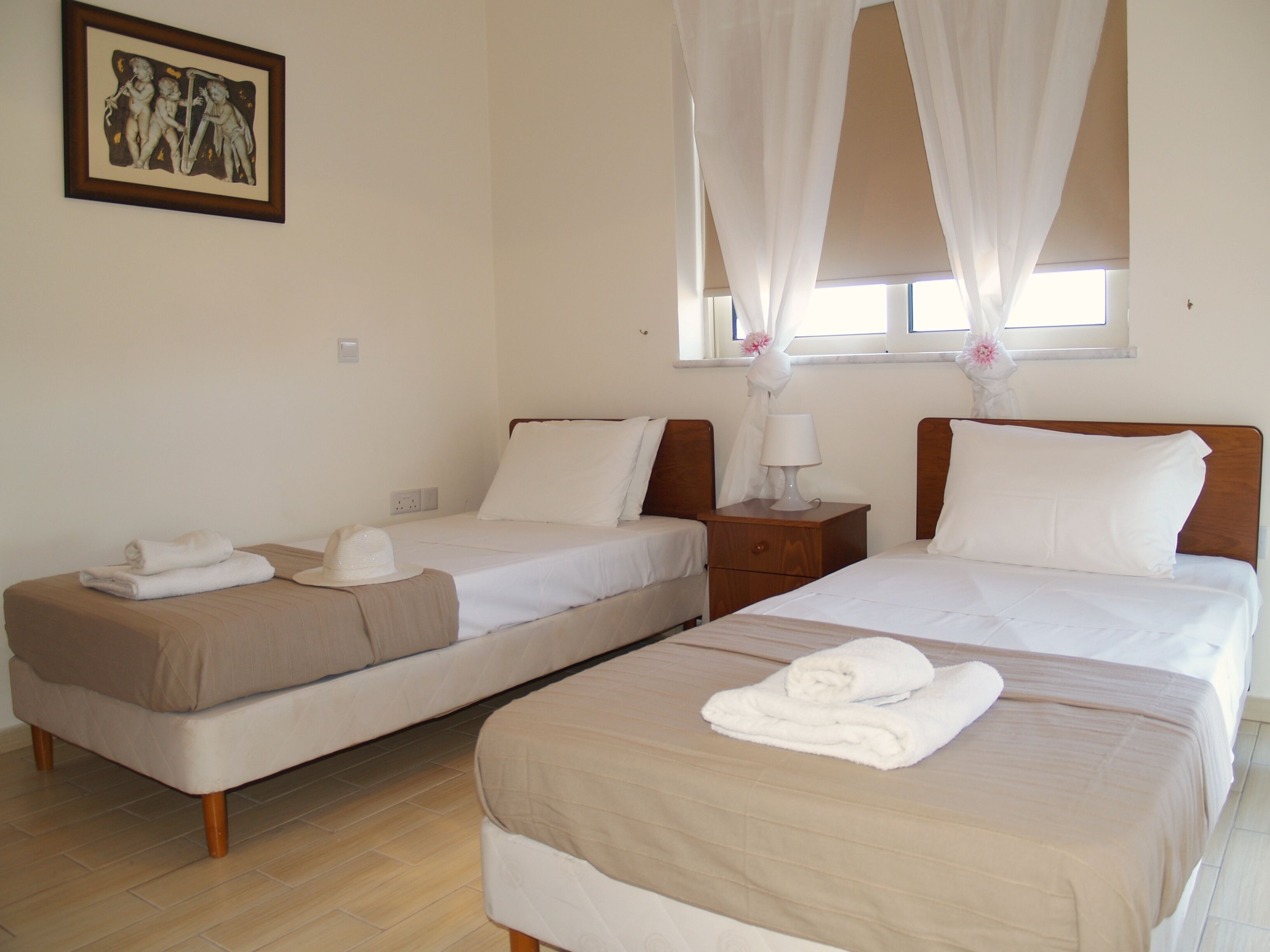 A hotel room with two beds, a nightstand, a bedside table with a lamp, a framed picture on the wall, and a window with sheer curtains.