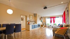 Een ruime woonkamer met een eettafel en stoelen, een keukenhoek, en een bank met kussens.