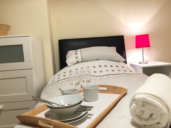 A neatly made bed with heart-patterned bedding, a bedside table with a lamp, and a tray with bowls, spoons, and a cup.