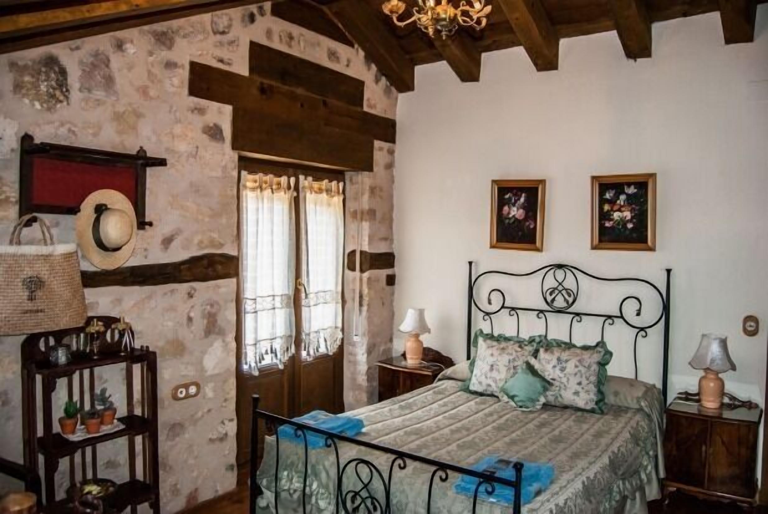 Ein rustikales Schlafzimmer mit einer Steinwand, Holzbalken, einem Bett mit Metallgestell, zwei Nachttischen mit Lampen und einem an der Wand befestigten Hutfänger.