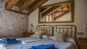 A bedroom with two beds, wooden beams, stone walls, a large mirror, and a nightstand with a lamp.