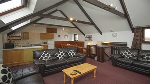 A cozy living room with two leather sofas, a wooden coffee table, and a high ceiling with exposed beams. A kitchen is visible in the background with wooden cabinets and a dining area.