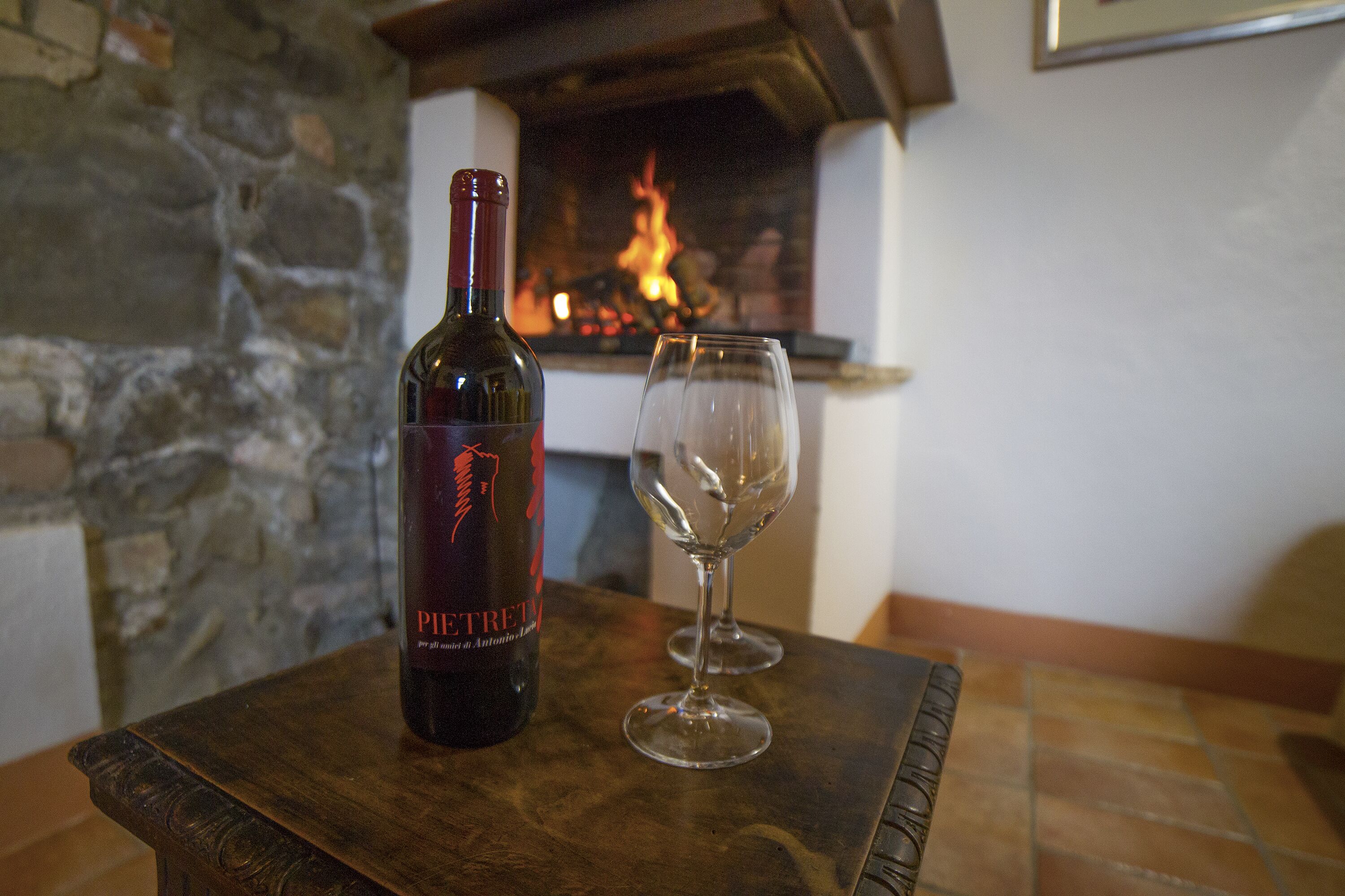 Una botella de vino y una copa vacía sobre una mesa de madera en una habitación con pared de piedra y una chimenea al fondo.