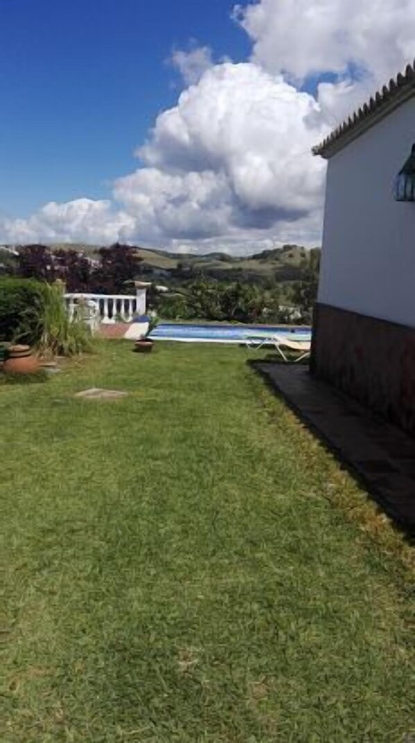 Un césped con piscina, un edificio blanco y colinas al fondo.