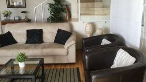 Ein Wohnzimmer mit einem beigefarbenen Sofa, einem schwarzen Ledersessel, einem Glastisch und einer Treppe im Hintergrund.