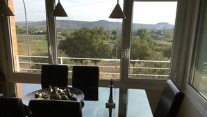 Ein Essbereich mit Tisch und Stühlen, einer Vase und Blick auf eine ländliche Landschaft durch große Fenster.