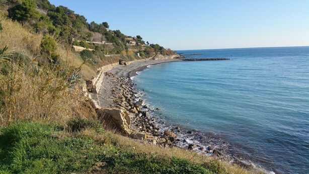 Paesaggio costiero con una costa rocciosa, cielo sereno e vegetazione su una collina.