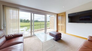 A spacious living room with a large sliding glass door leading to a patio, a leather sofa, a glass coffee table, and a flat-screen TV mounted on the wall. - Meadow View | Witney Holiday Let | 1 Bed & Sleeps 2 (Witney)