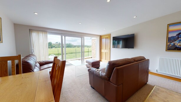 A spacious living room with a brown leather sofa, a wooden dining table, and a large sliding glass door leading to a garden. - Meadow View | Witney Holiday Let | 1 Bed & Sleeps 2 (Witney)