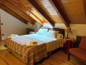 Ein gemütliches Schlafzimmer im Dachgeschoss mit Holzbalkendecke, einem Bett mit Karo-Bettwäsche, zwei Nachttischlampen und einem roten Sessel.