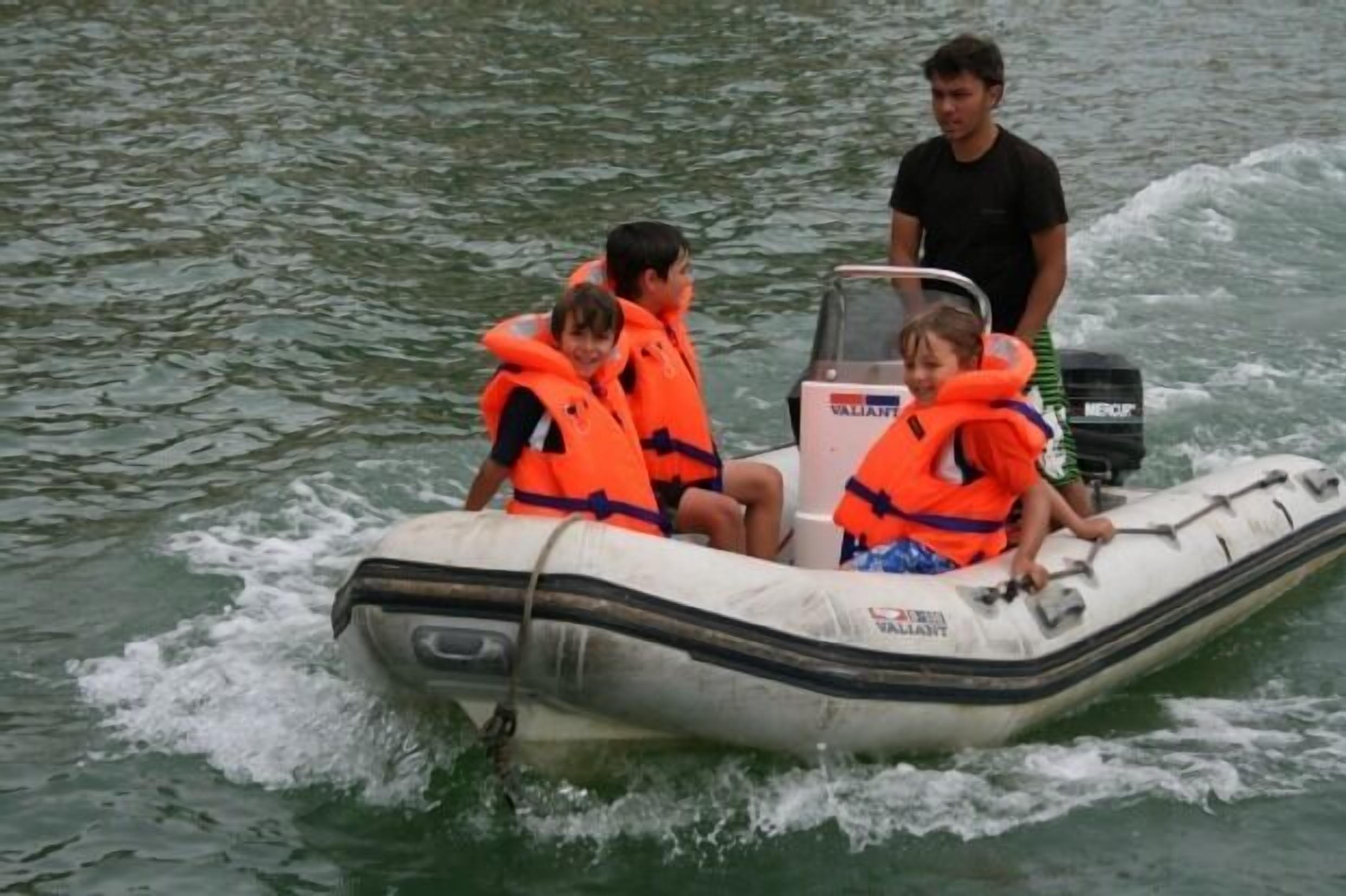 Ein kleines Schlauchboot mit drei Personen in Schwimmwesten, eine Person steht im Heck, und ein Motor mit der Aufschrift "VALIANT" ist an der Seite angebracht.
