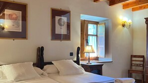 A room with two beds, wooden ceiling beams, framed artwork, a window with curtains, and a nightstand with a lamp. - The Abode of Vadinia (Burón)