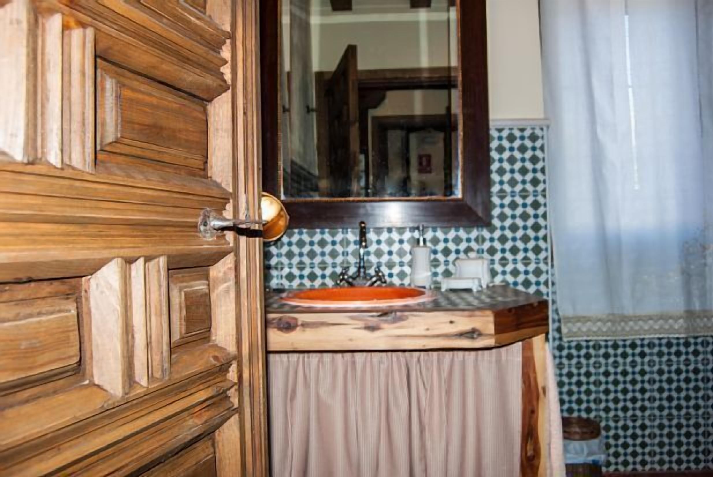 Una puerta de madera con un picaporte de latón, un espejo sobre un lavamanos con encimera de madera y salpicadero de azulejos estampados.