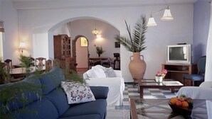 Un soggiorno con un divano blu, una poltrona bianca, un tavolino da caffè con una ciotola di frutta, un televisore e un vaso grande con fiori.