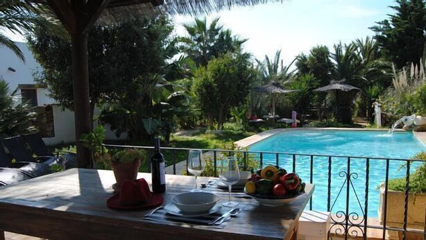 Un tavolo di legno con una ciotola di frutta, una bottiglia di vino e bicchieri su un balcone con vista su una piscina.