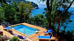 Une terrasse en bois avec une piscine et des chaises longues, donnant sur une zone côtière bordée d’arbres et offrant une vue sur la mer.