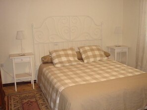 Una cama con colcha a cuadros, dos almohadas y un cabecero de metal blanco. Hay dos mesitas de noche, cada una con una lámpara. Un estante pequeño está adosado a la mesita de la izquierda.