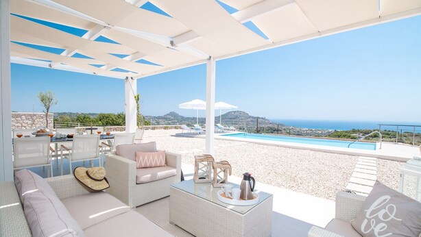 Zona relax all'aperto illuminata dal sole, con piscina, mobili in vimini bianco e vista libera sul mare.
