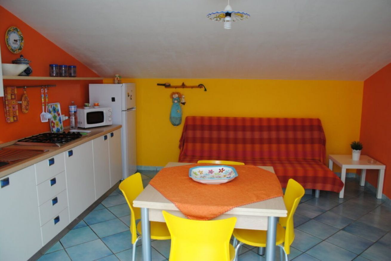 Une cuisine compacte équipée d’un four à micro-ondes, d’une cuisinière et de placards. On y trouve une table à manger avec des chaises jaunes et un canapé rouge dans l’espace repas.