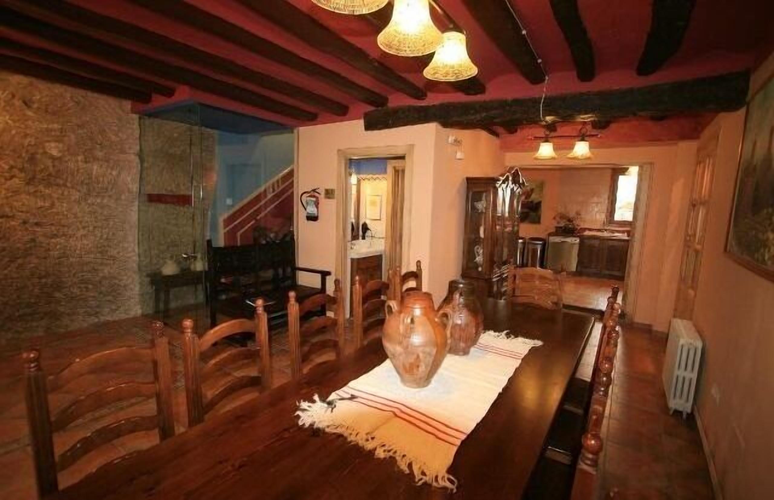 A dining area with a long wooden table, chairs, and a central vase. There is a staircase in the background leading to another room.