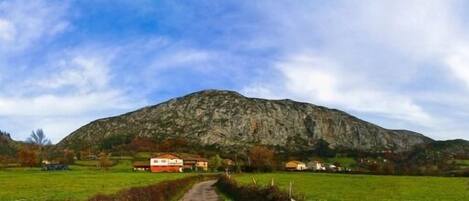 A rural landscape with a prominent mountain, a path leading to a cluster of houses, and a clear sky.