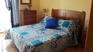 Een slaapkamer met een houten bed, een blauw kussen, een bedovertrek met patroon, een houten dressoir, een lamp en een ingelijst schilderij aan de muur.