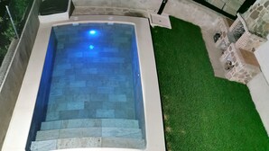 Una piscina con interior iluminado en azul, rodeada por un muro de piedra y una zona cubierta de césped.