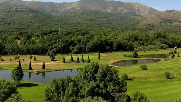 Un campo de golf con un estanque, árboles y montañas al fondo.